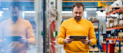 A man wearing a yellow sweater is looking at a tablet in an industrial setting background.