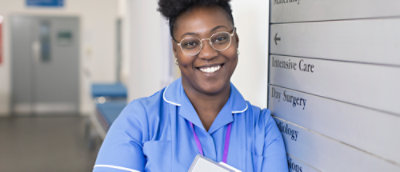A woman wearing glasses and a blue shirt, smiling at the camera with a sign in the background reading Intensive Care Day Surgery.