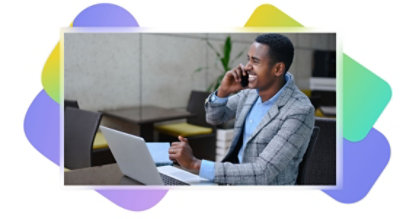 Eine Person, die mit einem Laptop an einem Tisch sitzt, ein Mobiltelefon ans Ohr hält und lächelt