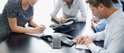 Trzy osoby siedzą przy stole podczas spotkania i sporządzają notatki. Na stole znajdują się telefony konferencyjne.