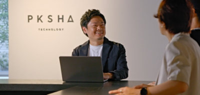 A person sitting at a table with a laptop displaying text that reads PKSHA.