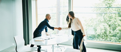 שני אנשים בחדר ישיבות, עומדים ולוחצים ידיים מעל שולחן עגול ועליו דפים ומחשב נישא