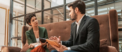 Deux individus assis sur un canapé discutent, l'un d'eux tenant une tablette numérique.