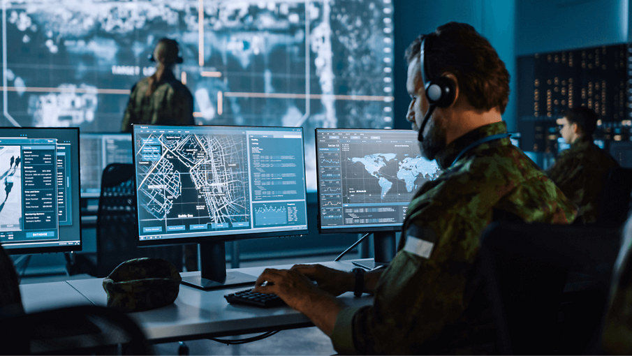 A member of the military wearing a headset working at a desk viewing maps and city grids