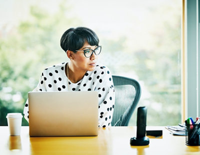 A person wearing glasses and a polka dot shirt sits at a desk with a laptop, looking to the side.