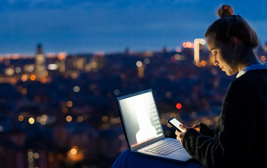 Woman using a cell phone and laptop at dawn overlooking a city.