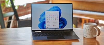 Een laptop op een houten tafel, met een Windows 11-bureaublad met geopende apps. Rechts staat een gespikkelde mok.