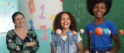 Zwei lächelnde Kinder, die mit einer Lehrerin vor einem Planetenmodell in einem Klassenzimmer stehen