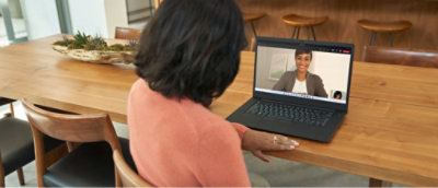 A woman sits at a wooden table, engaging in a video call on her laptop with another woman who is smiling on the screen.