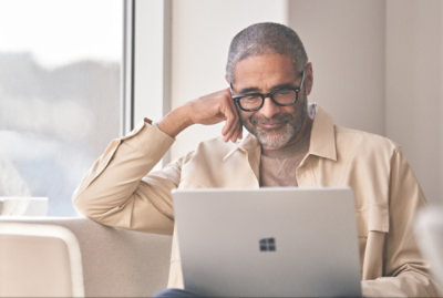 A man wearing glasses and a beige jacket is sitting on a couch, looking at a laptop screen and smiling.