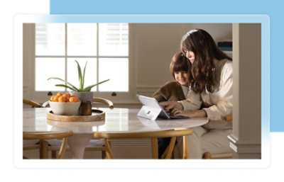 A woman and a child sit at a round table, looking at a tablet. A bowl of oranges and a potted plant are on the table.