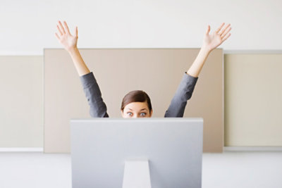 Osoba sedi iza monitora računara sa podignutim rukama, kao da slavi ili pokušava nešto da dohvati. 