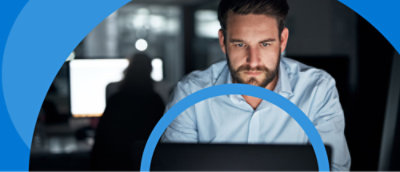 A man with a beard looks intently at a laptop screen in a dimly lit office, with a blue graphic overlay partially encircling him.