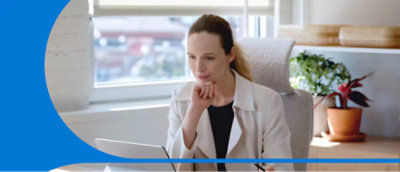 A person sits at a desk, attentively looking at a laptop screen, with a pen in hand. 
