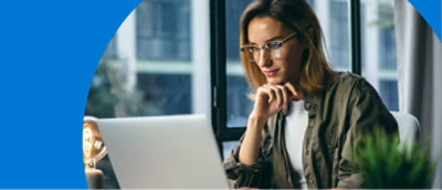 Person wearing glasses and a green jacket sits at a desk, looking at an open laptop with a focused expression.