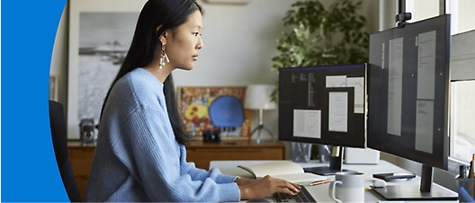 A woman in a blue sweater works at a dual-monitor setup in a home office with plants and artwork in the background.