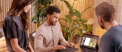  мужчина сидит за столом с ноутбуком, а два других человека стоят.