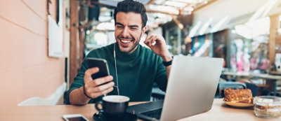 Uśmiechnięty mężczyzna w zielonym swetrze siedzi przy stole i korzysta ze smartfona. Przed nim leży laptop, filiżanka kawy i notatnik. Ma założone słuchawki.