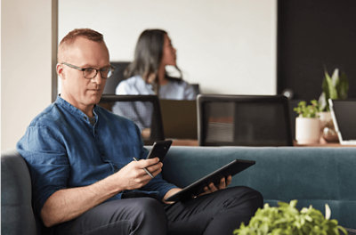 Een persoon die zijn telefoon gebruikt terwijl hij in een andere hand een tablet vasthoudt terwijl hij op de bank zit