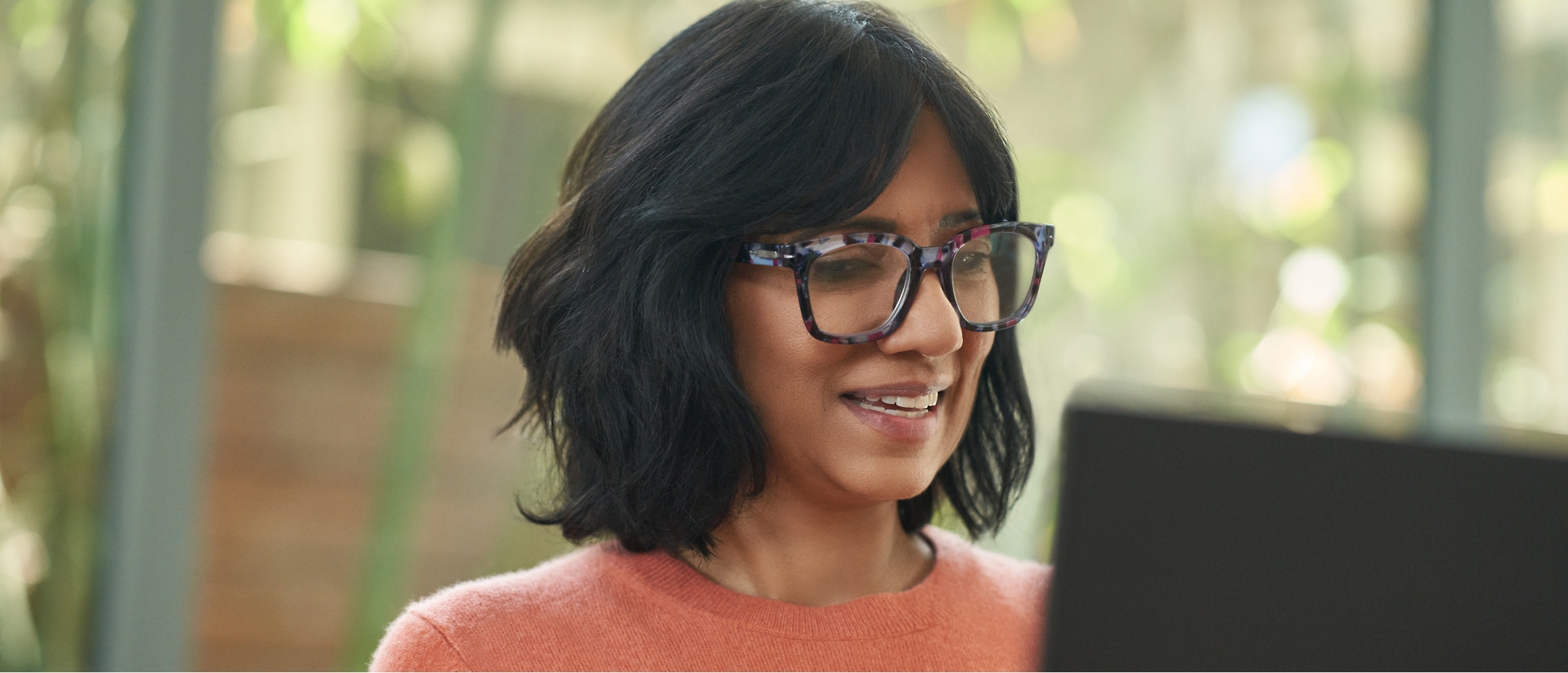 A person with short black hair and glasses is smiling while looking at a laptop screen. They are wearing an orange sweater.