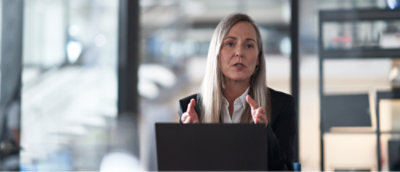 A person with long hair and a serious expression sits at a desk with a laptop, gesturing with both hands, in a modern office setting.