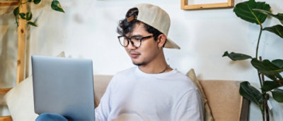 A man wearing a hat and glasses sitting on a couch using a laptop.