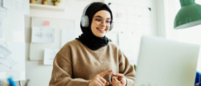 Жена със слушалки и хиджаб, която се усмихва докато гледа лаптопа си в осветена стая. 