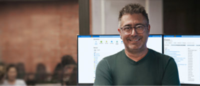 A person wearing spectacles smiling with a desktop screen in the background.