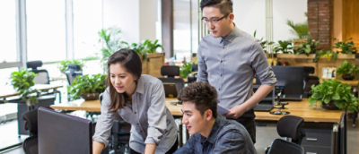 Трима души, които работят заедно на бюро с компютри в модерно офисно пространство с растения и естествена светлина.