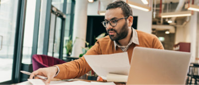 Un homme, dans un bureau, portant des lunettes et un pull orange, regarde des papiers qu'il tient d'une main tout en utilisant un ordinateur portable.