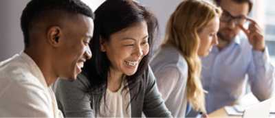 Quatre personnes sont assises à une table et discutent en travaillant sur des ordinateurs portables. Elles semblent être dans un contexte collaboratif.