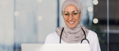 A woman in a white coat holding a phone and looking at a computer screen.