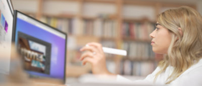 Frau in einer hellen Büroumgebung, die einen Tablettstift verwendet, um mit einem Grafikdesign zu interagieren, das auf einem Computermonitor angezeigt wird.