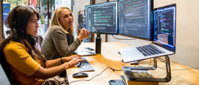 Zwei Frauen, die an einem Schreibtisch zusammenarbeiten, mit mehreren Computerbildschirmen, die Code anzeigen.