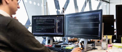 Ein Programmierer, der in einer Büroumgebung an mehreren Monitoren arbeitet, auf denen Code angezeigt wird.