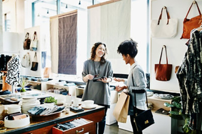 Dve žene razgovaraju u maloprodajnoj prodavnici, jedna drži telefon a druga nosi torbe za kupovinu. 