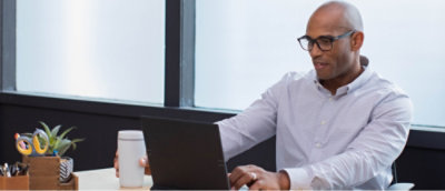 Person in a white shirt working on a laptop at an office desk with a coffee cup and plant pot.