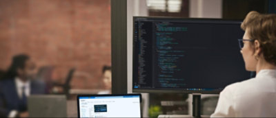 Osoba pracuje na počítači v kanceláři, přičemž na jednom monitoru má kód a na druhém e-maily.