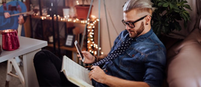 Mężczyzna w okularach i siwym kucyku trzyma smartfon i patrzy na niego, siedząc i pisząc w notatniku. Ma na sobie niebieską koszulę i wzorzysty krawat.