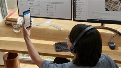 A person sitting at their desk wearing a headset and lifting up their mobile phone to look at their calendar