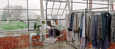 Muškarac u svetloplavoj košulji radi na laptop računaru u prodavnici odeće u kojoj razni odevni predmeti vise na šinama za kačenje odeće