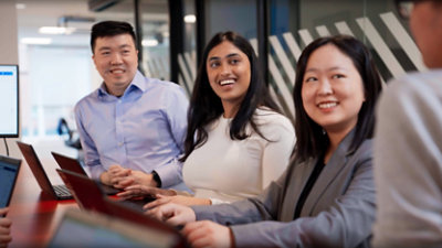 Four people in business attire are sitting at a conference table with laptops, engaging in conversation and smiling.