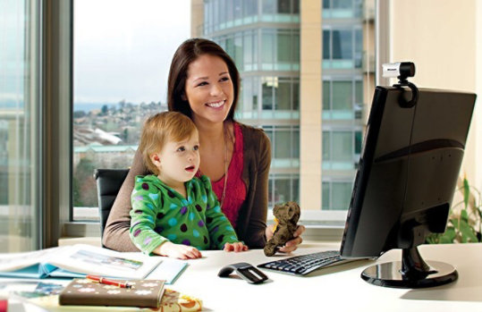 Uma mulher com uma criança no colo, sentada em uma mesa olhando para um monitor com uma webcam LifeCam Studio encaixada nele