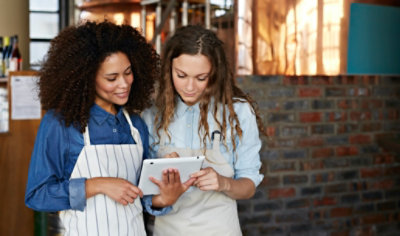 Dve žene sa keceljama stoje zajedno i gledaju tablet. Jedna žena drži tablet, a druga pokazuje na ekran. 