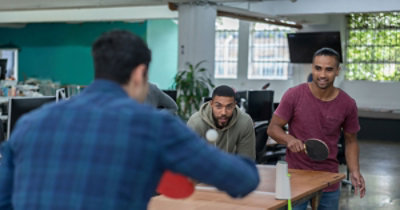 Dvojica muškaraca igraju stoni tenis u zatvorenom prostoru, a ostali ih gledaju.