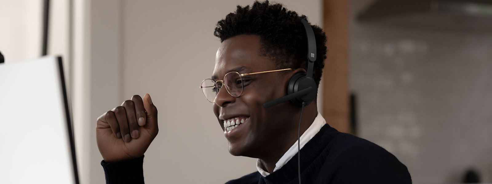 A male wearing a Microsoft Modern USB-C Headset, smiles at his computer screen with his hand raised