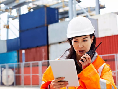 A ground staff wearing safety jacket holding tablet and talking using walkie talkie.