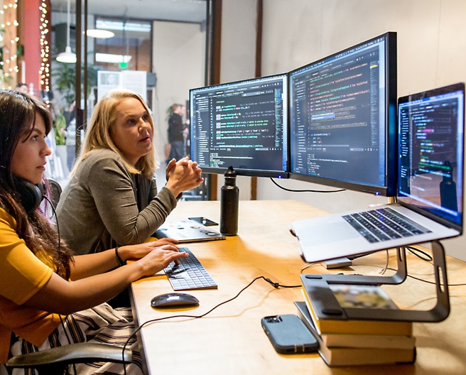 Zwei Frauen, die an einem Schreibtisch mit mehreren Monitoren arbeiten und sich unterhalten
