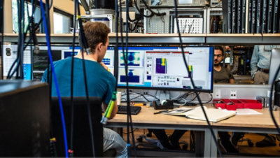 A person working at their desk with multiple screens in a room filled with wires