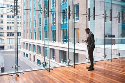 Человек, стоящий перед стеной окон и пользующийся мобильным телефоном.